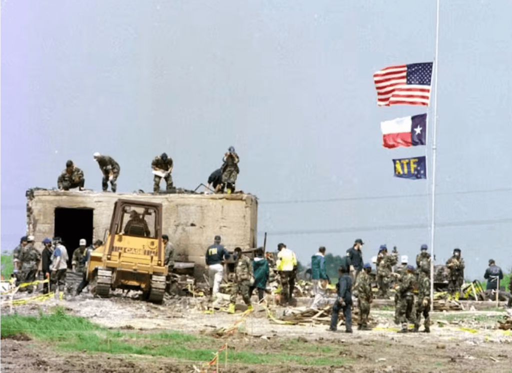 FBI agents search the site of the burned down building. The concrete vault and its open doorway are visible, along with flags raised by ATF agents. Waco Herald-Tribune